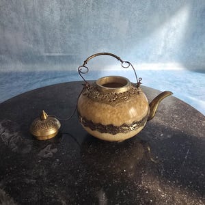Könnte beinhalten: Eine runde Teekanne mit einem beigen, marmorierten Steinkörper, der mit verzierten, dunklen Metallfiligranbändern am Hals und in der Mitte akzentuiert ist. Sie verfügt über einen geschwungenen Metallausguss, einen dünnen Drahthenkel und einen passenden Metalldeckel.