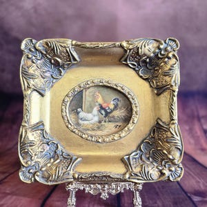 Peut inclure: Un petit cadre doré orné d'une peinture miniature d'un coq et d'une poule. Le cadre présente des bords sculptés et détaillés et repose sur un support en métal décoratif. La peinture représente un coq aux plumes rouges, noires et jaunes.