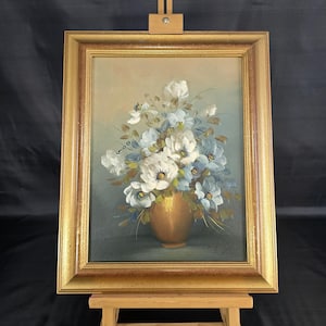 Peut inclure: Peinture à l'huile de fleurs blanches et bleues dans un vase doré, sur fond neutre. L'œuvre est encadrée dans un cadre en bois doré et exposée sur un petit chevalet en bois. Le style de la peinture rappelle la nature morte florale classique.