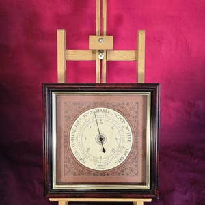 May include: A square barometer in a dark wood frame with a gold inner border. The cream-colored dial features black text including "RAIN," "VARIABLE," "SCHON," "FINE," "VERY DRY," and "STURM," with a central needle and a secondary adjustment hand.