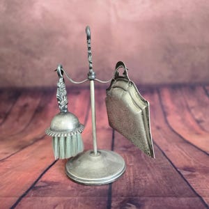 Peut inclure: Un support en métal argenté composé d'une tige verticale centrale sur une base circulaire. Suspendus aux bras incurvés supérieurs se trouvent une petite brosse à main aux poils clairs et une pelle à poussière assortie à surface texturée.