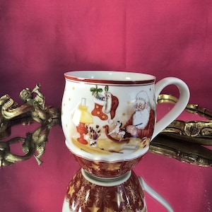 May include: A white ceramic Christmas mug with a red rim and handle. The mug features a festive illustration of Santa Claus, a stocking, and a lamp. The base of the mug has a red and gold star pattern.