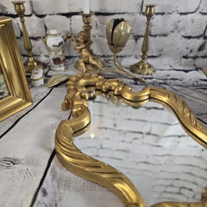 Peut inclure: Un miroir doré orné avec un cadre décoratif. L'image montre également plusieurs chandeliers dorés, une petite statue de chérubin et un bougeoir décoratif en forme de tulipe. Les objets sont disposés sur une surface blanche et grise avec un fond en briques.