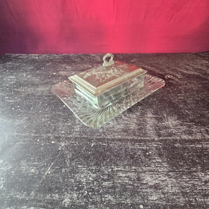 Peut inclure: Un beurrier rectangulaire composé d'une base en verre transparent avec un motif texturé en éventail et d'un couvercle amovible en métal argenté. Le couvercle est orné d'un motif floral gravé et surmonté d'une petite poignée en forme de boucle.