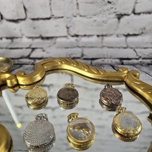 Puede incluir: Una colección de pequeños y ornamentados relojes de bolsillo en varios acabados metálicos, incluyendo oro, plata y bronce. Algunos relojes presentan diseños y grabados intrincados, mientras que otros tienen esferas claras. Los relojes se exhiben sobre una superficie reflectante.