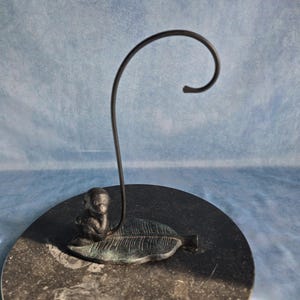 Op de afbeelding: Een donkere metalen displaystandaard met een klein apenfiguurtje op een voet in de vorm van een gestructureerd blad. Een dunne, gebogen metalen haak steekt omhoog uit het blad, bedoeld voor het ophangen van kleine voorwerpen met een donkere afwerking.