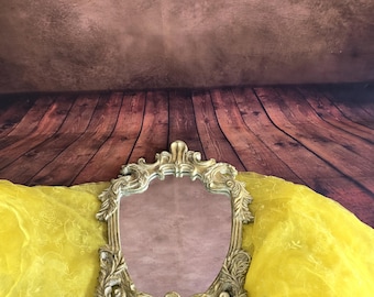 May include: A decorative, ornate gold-colored mirror with a detailed frame. The mirror is set against a backdrop of yellow fabric and a brown wooden surface.