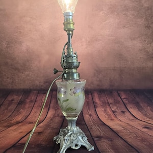 Puede incluir: Una lámpara de mesa antigua con una pantalla de cristal esmerilado adornada con diseños florales morados. La lámpara presenta una base plateada y una sección superior de color latón, coronada por una bombilla encendida. La lámpara está sobre una superficie de madera.