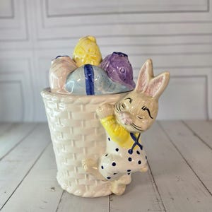 Peut inclure: Un panier de Pâques en céramique avec une figurine de lapin. Le panier est blanc avec une texture tissée et contient des objets colorés en forme d'œuf. Le lapin est crème avec des pois bleus et une chemise jaune. Le lapin est appuyé contre le panier.