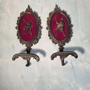 Peut inclure: Deux appliques murales antiques de couleur bronze. Chaque applique présente un cadre ovale avec un fond en velours bordeaux. L'une affiche un arc et une flèche, l'autre un casque et une lance. Les appliques ont des volutes décoratives et un crochet.