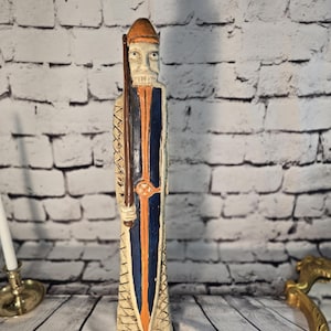 Könnte beinhalten: Eine geschnitzte Holzfigur eines Kriegers, der mit Schild und Schwert aufrecht steht. Die Figur hat einen Helm und ist mit orangefarbenen, blauen und braunen Details bemalt. Die Statue ist etwa 30 cm hoch.