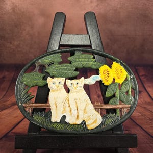 Könnte beinhalten: Ovales Zierelement aus Gusseisen mit zwei cremefarbenen Katzen, die nebeneinander sitzen. Die Katzen sind vor grünem Laub, einer gelben Sonnenblume und einem kleinen Holzzaun platziert. Das Ornament ist auf einer schwarzen Staffelei montiert.