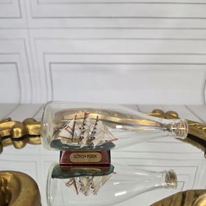 Puede incluir: Una botella de vidrio transparente que contiene un modelo de velero en miniatura. El modelo, llamado "Gorch Fock", presenta velas blancas y detalles rojos. La botella descansa sobre una superficie reflectante.