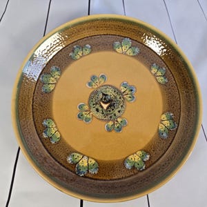May include: A round ceramic plate with a yellow center and a brown border. The plate features a decorative pattern of green and yellow leaf-like shapes. The rim is a darker brown with a textured appearance.