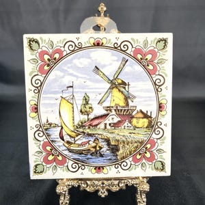 Peut inclure: Carreau de céramique carré avec une scène de moulin à vent hollandais, avec un voilier sur une rivière, un moulin à vent et un village. La tuile a une bordure florale rouge, rose et verte. Affiché sur un support doré décoratif.