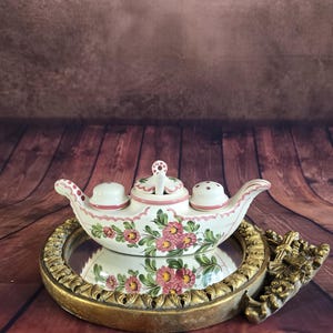 Peut inclure: Un ensemble de condiments en céramique blanche avec un motif floral rose et vert. L'ensemble comprend un récipient central avec un couvercle et deux récipients plus petits avec des couvercles. L'ensemble est présenté sur un miroir rond encadré d'or.