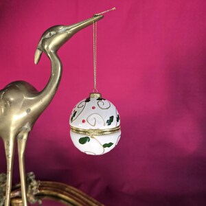 Peut inclure: Une boîte à bijoux ronde blanche, avec un bord doré et une chaîne dorée, décorée de feuilles de houx vertes et de baies rouges. La boîte est suspendue au bec d'une sculpture d'oiseau en laiton sur fond magenta.