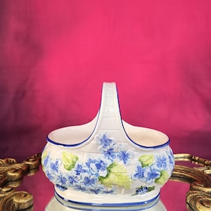 Peut inclure: Un panier en céramique blanc avec une anse, orné de fleurs d'hortensia bleues et de feuilles vertes. Le bord et la poignée sont soulignés en bleu. Le panier est posé sur une surface réfléchissante.