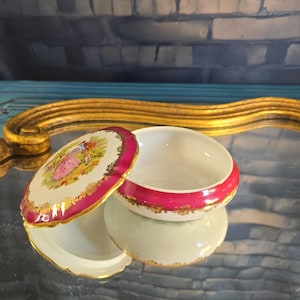May include: An open, ornate porcelain trinket box with a lid. The lid and rim are a vibrant magenta, accented with gold trim. The lid features a pastoral scene in the center. The box is reflected on a mirrored surface.