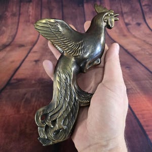 May include: A bronze-colored rooster figurine with detailed feathering on the wings and tail. The sculpture has a textured surface and is held in a hand, showcasing its size and craftsmanship. The rooster's head is upright, with a detailed comb and beak.