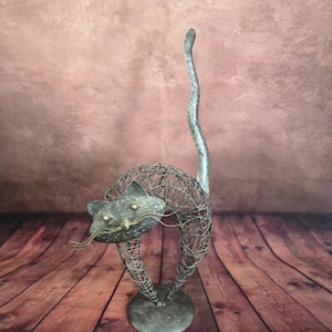 May include: A decorative cat sculpture crafted from dark grey wire mesh with a solid metal base. The cat features a flat, textured metal face with two small brass-colored eyes, a matching nose, and thin wire whiskers, topped with a long, upright tail.