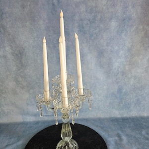 May include: A clear glass candelabra featuring four arms and a central stem, designed to hold five taper candles. The structure is adorned with hanging glass crystals and holds five slender white taper candles with pointed tips.