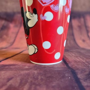 Puede incluir: Un vaso de cer&aacute;mica rojo con Mickey Mouse, lunares blancos y corazones rosas. El vaso tiene un borde y una base blancos. El dise&ntilde;o envuelve el vaso, mostrando la cara y el cuerpo de Mickey.