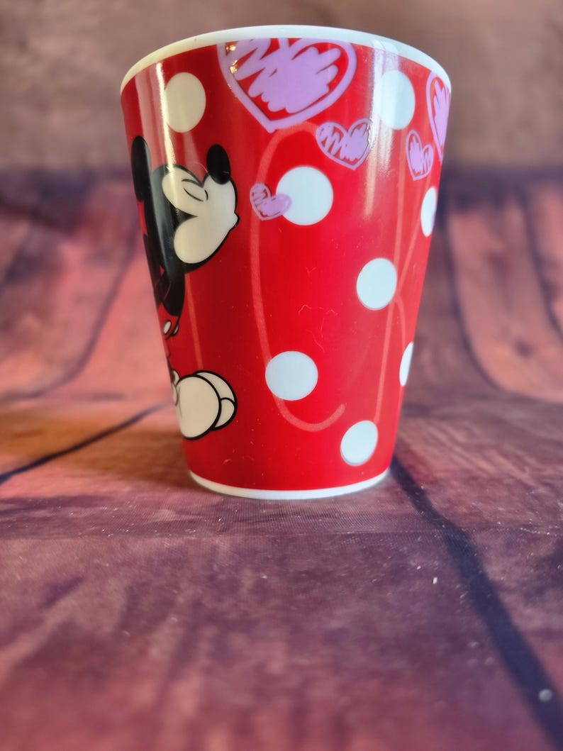 Puede incluir: Un vaso de cer&aacute;mica rojo con Mickey Mouse, lunares blancos y corazones rosas. El vaso tiene un borde y una base blancos. El dise&ntilde;o envuelve el vaso, mostrando la cara y el cuerpo de Mickey.