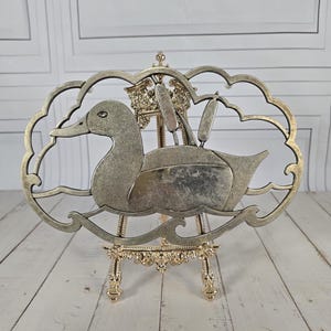 May include: A silver-toned metal duck sculpture with a textured surface, set within a cloud-shaped frame. The duck is depicted swimming, with cattails behind it. The sculpture is displayed on a decorative gold easel.