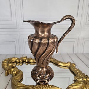 Può includere: Una brocca color rame con un design testurizzato e a spirale e un manico curvo. La brocca è appoggiata su un vassoio decorativo dorato e ornato. Lo sfondo è una parete chiara e decorata.