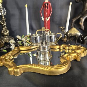 May include: A silver-toned metal condiment server featuring two small cups with handles, supported by a central vertical post with a top handle, all resting on a decorative gold-colored mirror tray with an ornate, scalloped frame edge.