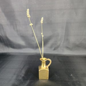 Könnte beinhalten: Eine kleine, goldfarbene Vase mit Henkel, die getrocknete Weizenstängel enthält. Die Vase hat einen quadratischen Boden und einen zylindrischen Hals. Die Weizenstängel sind hellbraun und die Vase ist aus Metall.