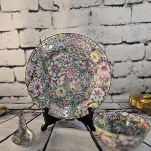 Peut inclure: Une assiette, un bol et une cuillère décoratifs avec un motif floral. L'assiette est soutenue par un support noir. L'assiette, le bol et la cuillère ont une bordure dorée. Le motif floral comprend des fleurs roses, violettes, jaunes et vertes.