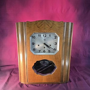 Peut inclure: Une horloge en bois vintage avec une finition marron clair. Le cadran de l'horloge est argenté avec des chiffres et des aiguilles noirs. L'horloge a un design géométrique décoratif et une section inférieure en miroir.