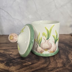 Puede incluir: Un tarro de ajo de cerámica blanca con tapa. El tarro cilíndrico tiene una base y un borde verdes. La tapa tiene un asa en forma de ajo. El tarro está decorado con ilustraciones de bulbos de ajo y hojas verdes.