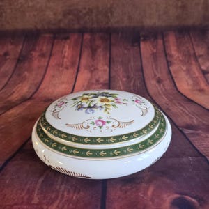 Peut inclure: Une boîte à bijoux ronde en céramique blanche avec un motif floral sur le couvercle. Le couvercle présente une bande verte avec un motif de flèches répétées. La boîte est ornée de fleurs roses, jaunes et violettes.