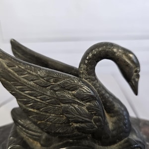 Peut inclure: Une sculpture détaillée de style antique représentant un cygne aux ailes déployées. Le cygne est d'une couleur métallique foncée, avec des détails de plumes complexes. La sculpture repose sur une base en marbre.