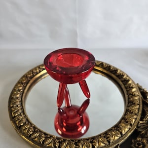 May include: A red glass candle holder with three legs. The candle holder is sitting on a gold mirror with a decorative frame.
