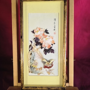 Puede incluir: Una pieza de arte mural rectangular con un diseño tridimensional de incrustaciones de conchas que muestra un ave con plumas de cola largas y flores rosas y blancas. La obra está sobre un fondo amarillo pálido en un marco de madera oscura.