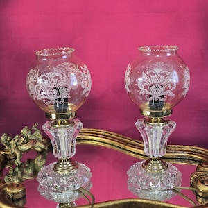 Peut inclure: Deux lampes de table vintage avec des abat-jours en verre transparent ornés d'un motif floral blanc. Les lampes ont une base dorée et une base en cristal clair facetté. Les lampes sont sur un miroir encadré d'or.