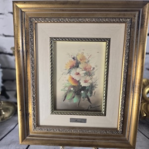 Peut inclure: Peinture florale encadrée représentant un bouquet de fleurs blanches, jaunes et oranges. La peinture est encadrée dans un cadre doré avec un passe-partout crème. Le cadre est soutenu par un support décoratif.