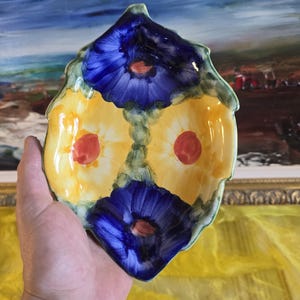 Peut inclure: Un plat en céramique peint à la main en forme de feuille, avec un motif floral. Le plat a une base jaune avec deux fleurs bleues et des accents rouges. Les bords sont verts.