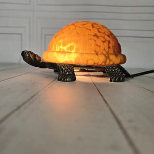 Könnte beinhalten: Schildkrötenförmige Lampe mit orangefarbener, strukturierter Schale. Der Sockel und der Kopf der Lampe sind dunkelgrün, und die Schale leuchtet, wenn sie eingeschaltet ist. Die Lampe steht auf einer hellen Holzoberfläche.