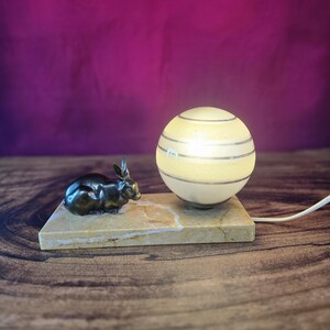 Peut inclure: Une lampe de table composée d'un abat-jour sphérique en verre dépoli à bandes horizontales, posée sur un socle rectangulaire en marbre beige à côté d'une petite figurine de lapin en bronze foncé. Un cordon d'alimentation blanc dépasse du socle.