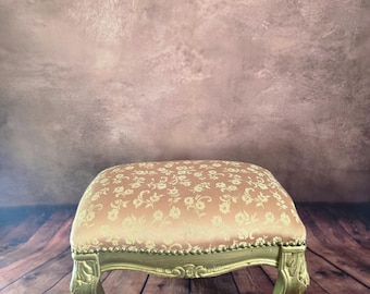 May include: A rectangular footstool featuring a dusty pink upholstered top with a cream-colored floral pattern. The base is constructed from gold-painted wood with carved cabriole legs and a row of decorative metal nailhead trim along the edge.