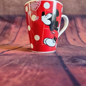 Puede incluir: Taza de cer&aacute;mica roja con Mickey Mouse en pose de beso, con lunares blancos y formas de coraz&oacute;n. La taza tiene un asa y borde blancos, y mide aproximadamente 10 cm de alto.