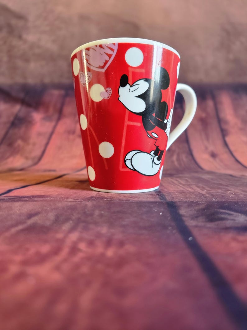 Puede incluir: Taza de cer&aacute;mica roja con Mickey Mouse en pose de beso, con lunares blancos y formas de coraz&oacute;n. La taza tiene un asa y borde blancos, y mide aproximadamente 10 cm de alto.