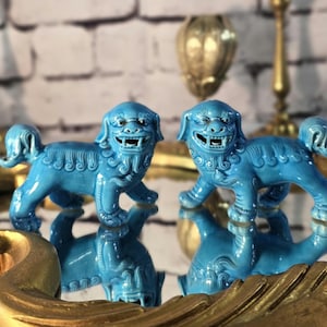 May include: Two turquoise ceramic Foo Dogs, also known as Chinese guardian lions, are displayed on a mirrored surface. The statues have detailed features and are reflected below. The background includes gold decorative elements.