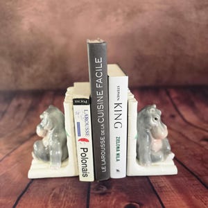Peut inclure: Une paire de serre-livres en céramique représentant des hippopotames gris assis sur des bases de style bambou crème. Les serre-livres soutiennent trois livres intitulés Larousse Polonais, Le Larousse de la Cuisine Facile et Stephen King Zielona Mila.