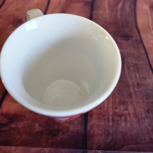 Puede incluir: Una taza de cer&aacute;mica blanca con un asa peque&ntilde;a. El interior de la taza es blanco y el borde tiene un sutil acento rosa. La taza est&aacute; sobre una superficie de madera marr&oacute;n.
